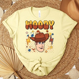 Puede incluir: Camiseta amarilla clara con un gr&aacute;fico de dibujos animados de Woody de Toy Story. La palabra "WOODY" est&aacute; impresa en letras de burbujas marrones y naranjas sobre la cara del personaje. El dise&ntilde;o incluye estrellas, rayos y un sombrero de vaquero marr&oacute;n.