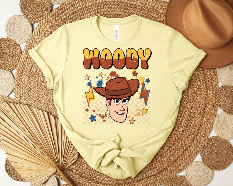 Puede incluir: Camiseta amarilla clara con un gr&aacute;fico de dibujos animados de Woody de Toy Story. La palabra "WOODY" est&aacute; impresa en letras de burbujas marrones y naranjas sobre la cara del personaje. El dise&ntilde;o incluye estrellas, rayos y un sombrero de vaquero marr&oacute;n.
