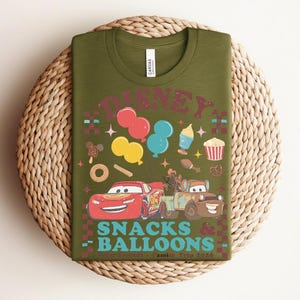 May include: Olive green t-shirt with a colorful graphic featuring the word "DISNEY" above cartoon images of balloons, snacks, and cars. The text "SNACKS & BALLOONS" is below the cars. The shirt is displayed on a woven mat.