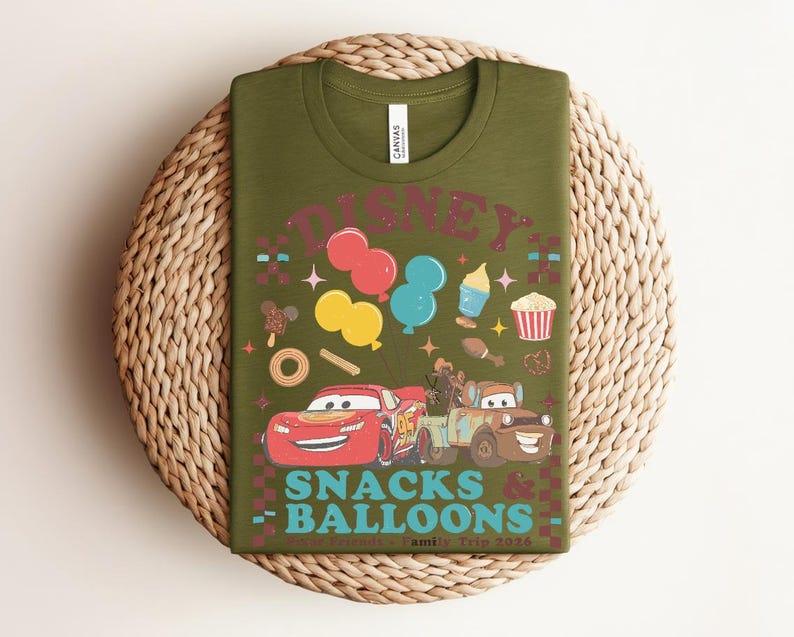 May include: Olive green t-shirt with a colorful graphic featuring the word "DISNEY" above cartoon images of balloons, snacks, and cars. The text "SNACKS & BALLOONS" is below the cars. The shirt is displayed on a woven mat.
