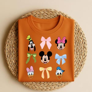 May include: An orange t-shirt with cartoon character faces and bows. The shirt features Goofy, Minnie Mouse, Mickey Mouse, Donald Duck, and Daisy Duck. The bows are in various colours, including pink, green, and blue.