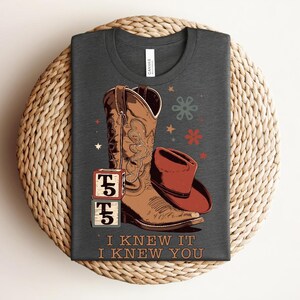 May include: A dark gray t-shirt featuring a graphic of a brown leather cowboy boot, a red cowboy hat, and two stacked blocks with the text T5. The shirt includes the text I KNEW IT I KNEW YOU below the graphic, along with small star and flower icons.