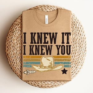 May include: A tan t-shirt featuring the black text I KNEW IT I KNEW YOU above a graphic of a cowboy hat, a small rocket, and a star. The design includes horizontal stripes in muted yellow, teal, and orange colors with a distressed texture.
