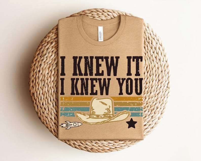 May include: A tan t-shirt featuring the black text I KNEW IT I KNEW YOU above a graphic of a cowboy hat, a small rocket, and a star. The design includes horizontal stripes in muted yellow, teal, and orange colors with a distressed texture.