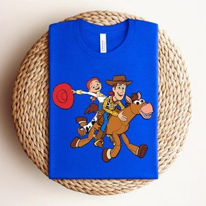 May include: A royal blue t-shirt featuring a colorful graphic of Woody, Jessie, and a horse from the animated film Toy Story. The design shows the characters riding the horse. The shirt is displayed on a woven, circular mat.