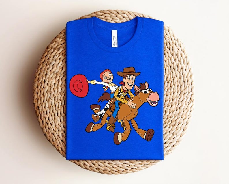 May include: A royal blue t-shirt featuring a colorful graphic of Woody, Jessie, and a horse from the animated film Toy Story. The design shows the characters riding the horse. The shirt is displayed on a woven, circular mat.
