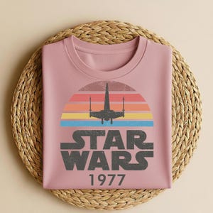 May include: A dusty rose-colored sweatshirt featuring a retro Star Wars design. The graphic includes a silhouette of a starfighter against a sunset of orange, yellow, and blue stripes, with the text "STAR WARS 1977" in gray.