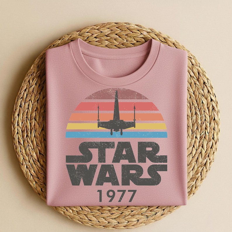 May include: A dusty rose-colored sweatshirt featuring a retro Star Wars design. The graphic includes a silhouette of a starfighter against a sunset of orange, yellow, and blue stripes, with the text "STAR WARS 1977" in gray.