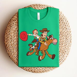 May include: Green t-shirt featuring a colorful graphic of Woody, Jessie, and a horse from the Toy Story movie. The design shows the characters riding the horse. The shirt is folded on a woven, circular surface.