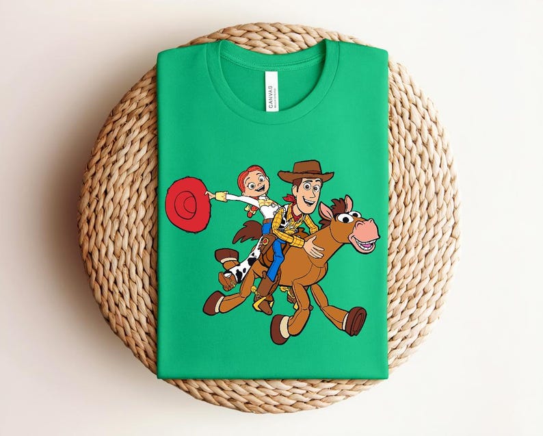May include: Green t-shirt featuring a colorful graphic of Woody, Jessie, and a horse from the Toy Story movie. The design shows the characters riding the horse. The shirt is folded on a woven, circular surface.