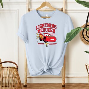 May include: Light blue t-shirt with a red cartoon race car and the text "Lightning McQueen" in red. The car is holding a trophy and the text "Piston Cup Racing Champion" is below the car.