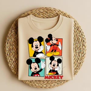 以下が含まれることがあります： ベージュのクルーネックTシャツ。ミッキーマウスのイラストが4つのパネルに描かれています。各パネルには、カラフルな正方形の中で異なるポーズのミッキーマウスが描かれています。「MICKEY」の文字が赤色で下部に印刷されています。