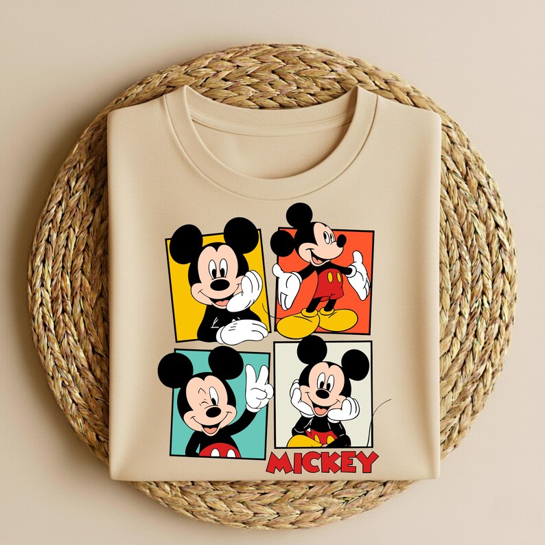 以下が含まれることがあります： ベージュのクルーネックTシャツ。ミッキーマウスのイラストが4つのパネルに描かれています。各パネルには、カラフルな正方形の中で異なるポーズのミッキーマウスが描かれています。「MICKEY」の文字が赤色で下部に印刷されています。