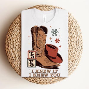 May include: A white t-shirt featuring a graphic of a brown cowboy boot, a red cowboy hat, two stacked alphabet blocks with the letter T, and small star and flower icons. The text "I KNEW IT I KNEW YOU" is printed in brown at the bottom.