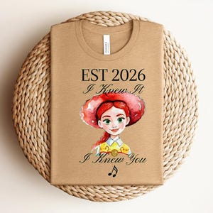 Pode incluir: Uma t-shirt bege com uma ilustra&ccedil;&atilde;o em estilo aguarela de uma personagem ruiva com um chap&eacute;u vermelho e camisa amarela. O texto diz EST 2026, I Knew It e I Knew You, acompanhado por um pequeno s&iacute;mbolo de nota musical preta abaixo.