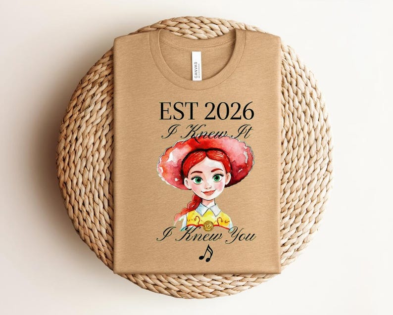 Pode incluir: Uma t-shirt bege com uma ilustra&ccedil;&atilde;o em estilo aguarela de uma personagem ruiva com um chap&eacute;u vermelho e camisa amarela. O texto diz EST 2026, I Knew It e I Knew You, acompanhado por um pequeno s&iacute;mbolo de nota musical preta abaixo.