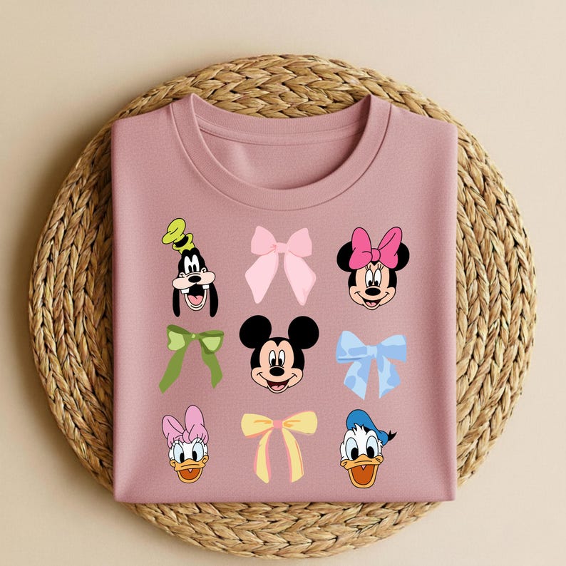 May include: A dusky pink sweatshirt with cartoon character faces and bows. Characters include Goofy, Mickey Mouse, Minnie Mouse, Donald Duck, and Daisy Duck. The bows are in various colours, including pink, green, and yellow.