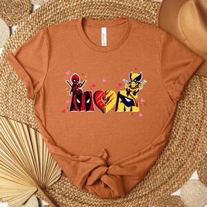 Op de afbeelding: Een roestkleurig t-shirt met een grafisch ontwerp met het woord "MOM" in een hartvorm, met Deadpool- en Wolverine-personages. Het ontwerp bevat roze hartjes en is gedrukt op een shirt met korte mouwen.
