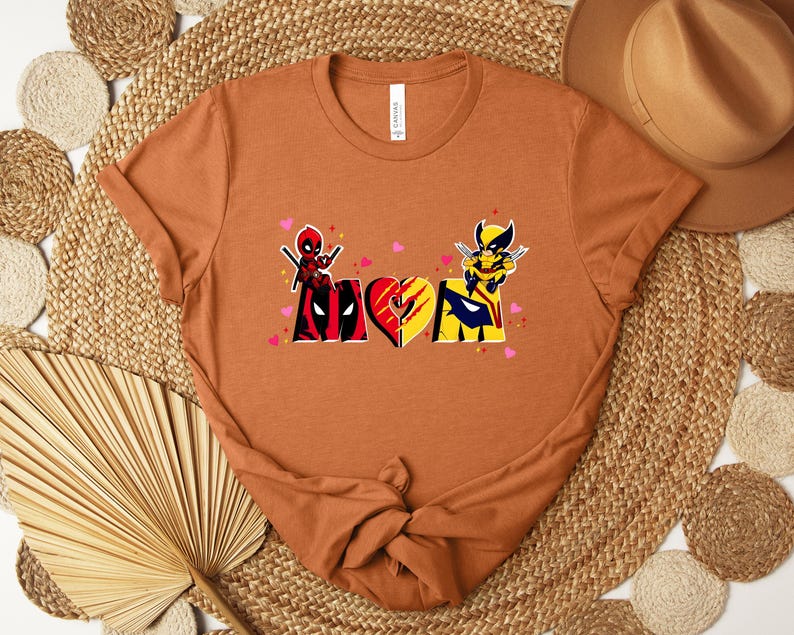 Op de afbeelding: Een roestkleurig t-shirt met een grafisch ontwerp met het woord "MOM" in een hartvorm, met Deadpool- en Wolverine-personages. Het ontwerp bevat roze hartjes en is gedrukt op een shirt met korte mouwen.