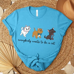 May include: A light blue t-shirt featuring three cartoon cats with the text "everybody wants to be a cat". The cats are white, brown, and black, each with unique accessories. The shirt is knotted at the bottom.