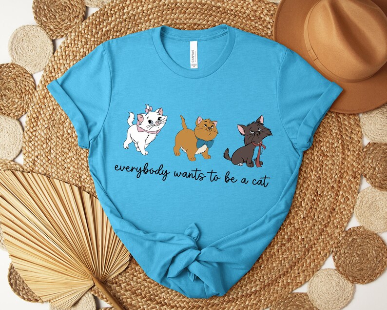 May include: A light blue t-shirt featuring three cartoon cats with the text "everybody wants to be a cat". The cats are white, brown, and black, each with unique accessories. The shirt is knotted at the bottom.