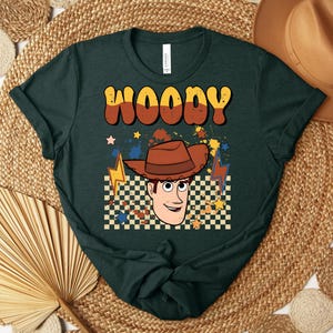 Puede incluir: Camiseta verde oscuro con la palabra "WOODY" en fuente retro naranja y marr&oacute;n. La camiseta tiene un gr&aacute;fico de un vaquero de dibujos animados con un sombrero marr&oacute;n, un fondo a cuadros y acentos de rayos y estrellas.