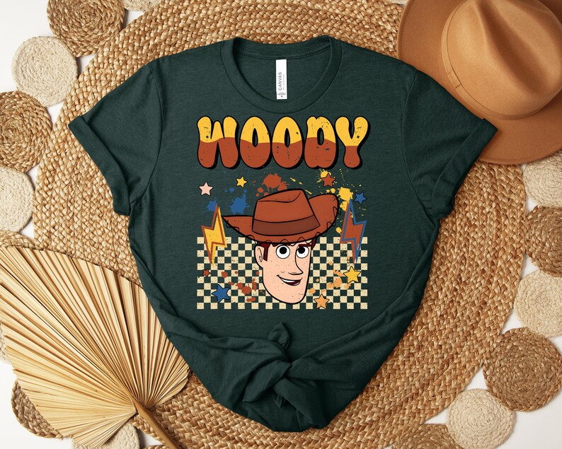 Puede incluir: Camiseta verde oscuro con la palabra "WOODY" en fuente retro naranja y marr&oacute;n. La camiseta tiene un gr&aacute;fico de un vaquero de dibujos animados con un sombrero marr&oacute;n, un fondo a cuadros y acentos de rayos y estrellas.