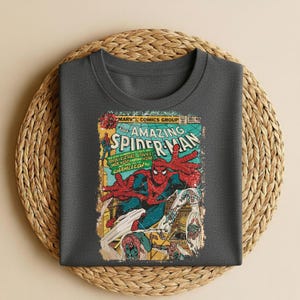 May include: A dark gray t-shirt featuring a vintage comic book cover of Spider-Man. The graphic includes the title "The Amazing Spider-Man" and other comic book elements. The shirt is displayed on a woven, circular mat.