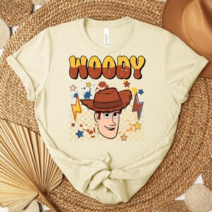 Puede incluir: Una camiseta de color beige claro con la palabra "WOODY" en fuente retro marr&oacute;n y amarilla. Debajo del texto hay una ilustraci&oacute;n de dibujos animados de un vaquero con un sombrero marr&oacute;n. El dise&ntilde;o incluye estrellas y rayos en azul, amarillo y naranja.