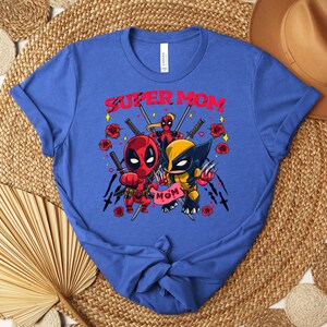 K&ouml;nnte beinhalten: Blaues T-Shirt mit einer Grafik von Deadpool und Wolverine und dem Text "SUPER MOM". Das Design umfasst Rosen, Schwerter und das Wort "MOM" auf einem Banner. Das T-Shirt hat einen Rundhalsausschnitt.