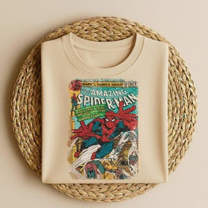 May include: A beige t-shirt featuring a vintage comic book cover of The Amazing Spider-Man. The comic art shows Spider-Man in a dynamic pose with red and blue costume, with text that reads "MARVEL COMICS GROUP" and "SPIDER-MAN".