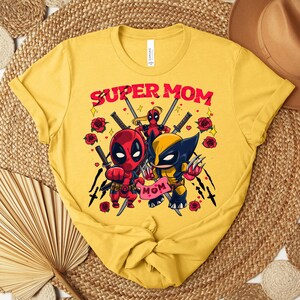 K&ouml;nnte beinhalten: Senfgelbes T-Shirt mit dem Text "SUPER MOM" in Rot, mit Cartoon-Illustrationen von Deadpool und Wolverine. Das Design umfasst Rosen und Schwerter und schafft ein Superhelden-Thema.