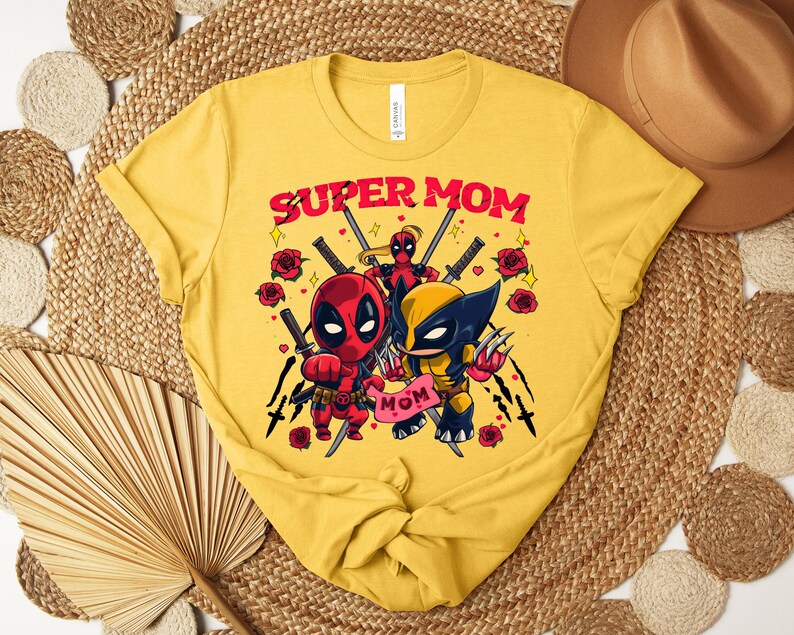 K&ouml;nnte beinhalten: Senfgelbes T-Shirt mit dem Text "SUPER MOM" in Rot, mit Cartoon-Illustrationen von Deadpool und Wolverine. Das Design umfasst Rosen und Schwerter und schafft ein Superhelden-Thema.