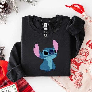 K&ouml;nnte beinhalten: Schwarzes Sweatshirt mit einem blauen Cartoon-Charakter mit gro&szlig;en Ohren. Eine silberne Halskette mit einem Sonnenanh&auml;nger ist sichtbar. Das Sweatshirt ist auf einer wei&szlig;en Oberfl&auml;che gefaltet.