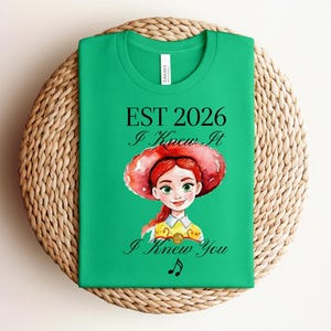 Pode incluir: Uma t-shirt verde brilhante de manga curta com uma ilustra&ccedil;&atilde;o em aguarela de uma personagem de cabelo ruivo e chap&eacute;u vermelho. O texto diz EST 2026, I Knew It e I Knew You, acompanhado por um pequeno s&iacute;mbolo de nota musical preta.