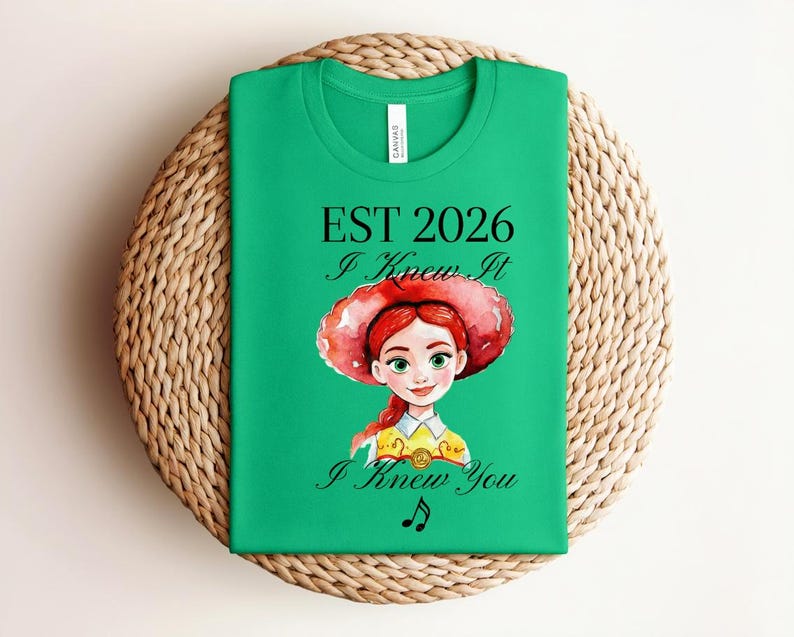 Pode incluir: Uma t-shirt verde brilhante de manga curta com uma ilustra&ccedil;&atilde;o em aguarela de uma personagem de cabelo ruivo e chap&eacute;u vermelho. O texto diz EST 2026, I Knew It e I Knew You, acompanhado por um pequeno s&iacute;mbolo de nota musical preta.