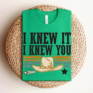 May include: A bright green t-shirt featuring the black text I KNEW IT I KNEW YOU above a graphic of a beige cowboy hat, a small rocket ship, and a black star, all set against horizontal stripes in muted orange, teal, and dark green colors.
