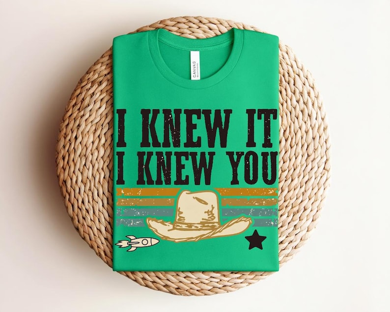 May include: A bright green t-shirt featuring the black text I KNEW IT I KNEW YOU above a graphic of a beige cowboy hat, a small rocket ship, and a black star, all set against horizontal stripes in muted orange, teal, and dark green colors.