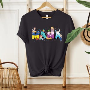 K&ouml;nnte beinhalten: Schwarzes T-Shirt mit einem Grafikdruck des Wortes "MAMA", das mit Disney-Prinzessinnen ausgeschrieben ist. Jeder Buchstabe ist eine andere Prinzessin, mit einem kleinen Bild der Prinzessin auf jedem Buchstaben.