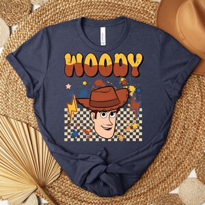 Puede incluir: Camiseta azul marino con la palabra "WOODY" en fuente retro marr&oacute;n y naranja. La camiseta presenta una ilustraci&oacute;n de dibujos animados de Woody con un sombrero de vaquero marr&oacute;n, sobre un fondo a cuadros con estrellas y rayos.