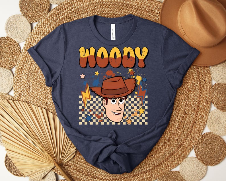 Puede incluir: Camiseta azul marino con la palabra "WOODY" en fuente retro marr&oacute;n y naranja. La camiseta presenta una ilustraci&oacute;n de dibujos animados de Woody con un sombrero de vaquero marr&oacute;n, sobre un fondo a cuadros con estrellas y rayos.