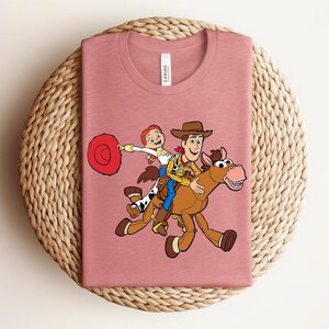 May include: A dusty rose t-shirt featuring a cartoon graphic of Woody, Jessie, and a horse. Woody is wearing a cowboy hat and a yellow shirt. Jessie is holding a red hat. The shirt is folded on a woven surface.