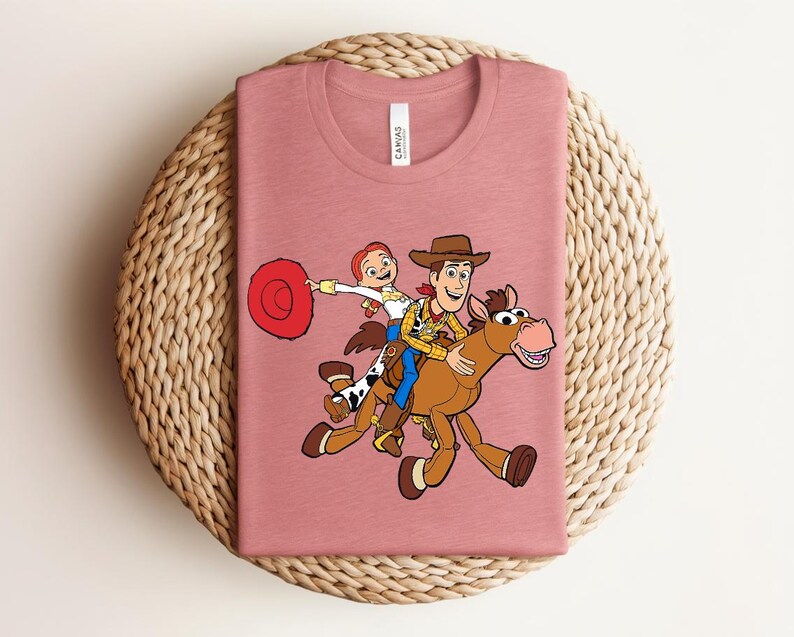 May include: A dusty rose t-shirt featuring a cartoon graphic of Woody, Jessie, and a horse. Woody is wearing a cowboy hat and a yellow shirt. Jessie is holding a red hat. The shirt is folded on a woven surface.