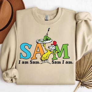 May include: Beige crewneck sweatshirt featuring the words "SAM" in blue, orange, and green, with the phrase "I am Sam. Sam I am." below. A cartoon character holds a plate of green eggs and ham. A red hat is on the character's head.