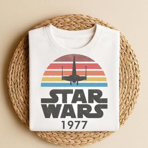 May include: White t-shirt featuring a retro design with a colorful sunset, a silhouette of a starfighter, and the text "STAR WARS 1977" in a distressed font. The shirt is displayed on a woven placemat.