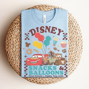 May include: Light blue t-shirt with a colorful graphic featuring the word "Disney" above a design of balloons, food, and cartoon cars. The text "Snacks & Balloons" and "Pixar Friends &bull; Family Trip 2026" are also printed on the shirt.
