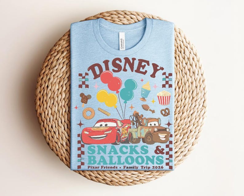May include: Light blue t-shirt with a colorful graphic featuring the word "Disney" above a design of balloons, food, and cartoon cars. The text "Snacks & Balloons" and "Pixar Friends &bull; Family Trip 2026" are also printed on the shirt.
