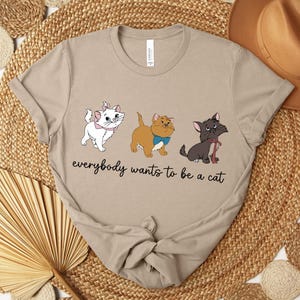 May include: Beige t-shirt featuring three cartoon cats with the text "everybody wants to be a cat". The cats are white, tan, and black, each with unique accessories. The shirt is displayed on a woven surface with a hat in the background.