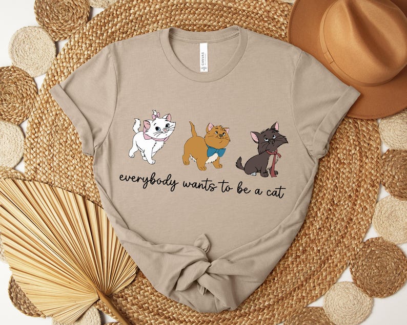 May include: Beige t-shirt featuring three cartoon cats with the text "everybody wants to be a cat". The cats are white, tan, and black, each with unique accessories. The shirt is displayed on a woven surface with a hat in the background.