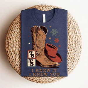 May include: A navy blue t-shirt featuring a graphic of a brown cowboy boot, a red cowboy hat, and two stacked alphabet blocks with the letter T. The design includes small stars and flower shapes, with the text I KNEW IT I KNEW YOU printed below.